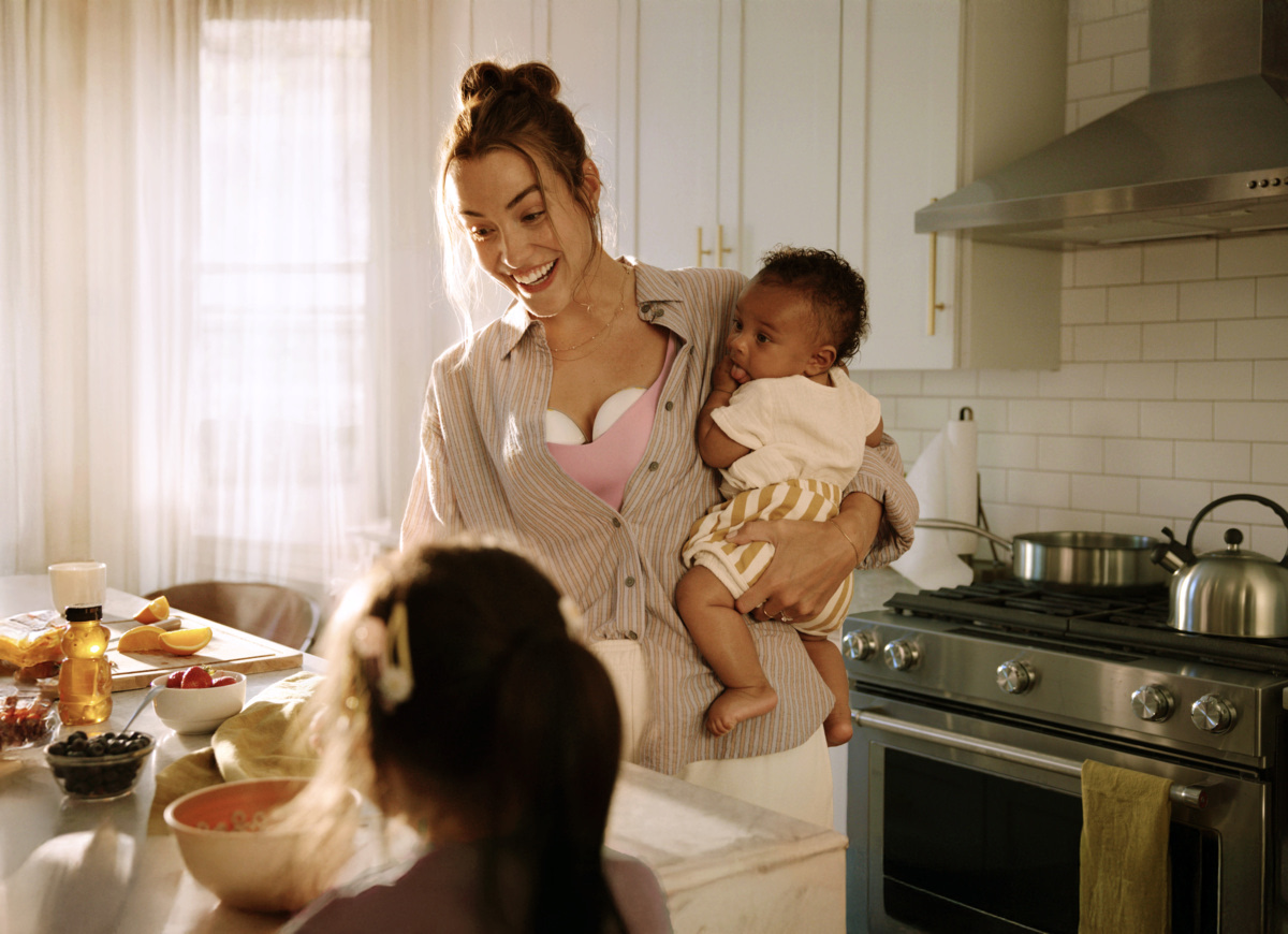 Maman tenant son bébé dans les bras tout en portant un tire-lait mains libres dans sa cuisine