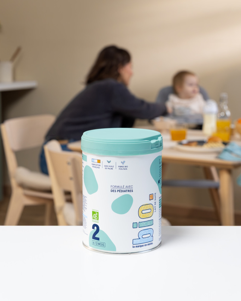 Boîte de lait infantile bio âge 2 la marque en moins posée sur une table, avec une maman et son bébé floutés à l'arrière-plan lors d'un repas