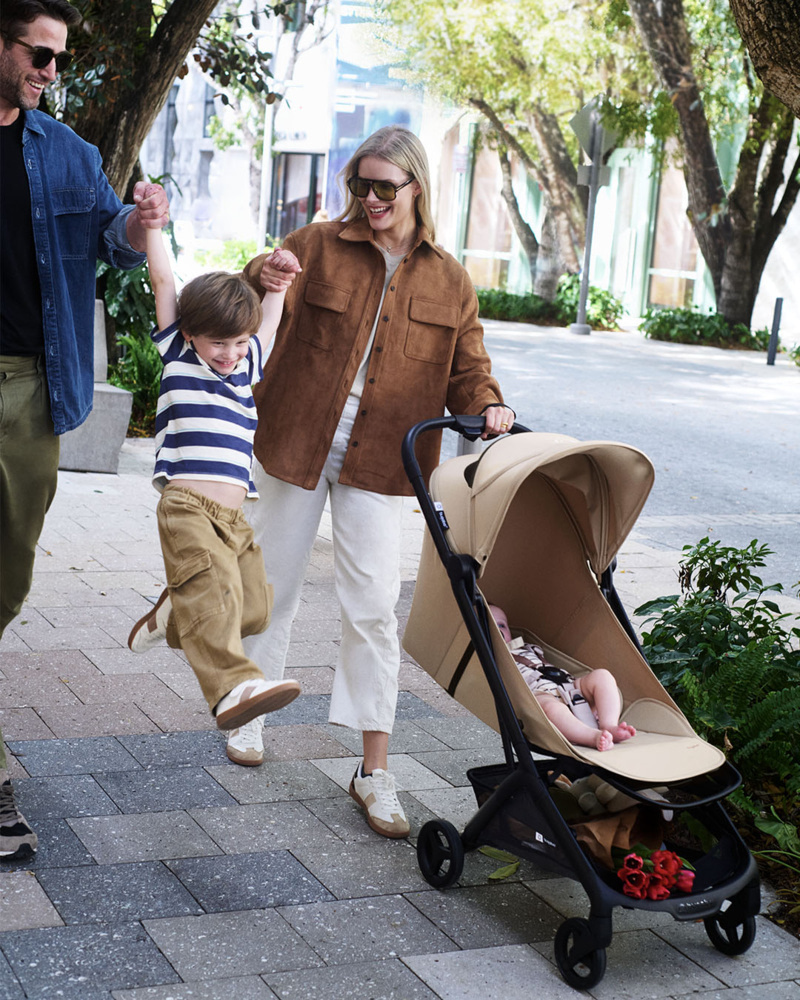 Une famille se promène en ville avec la poussette compacte Bugaboo Butterfly 2, un bébé confortablement installé et un enfant qui saute en tenant les mains de ses parents