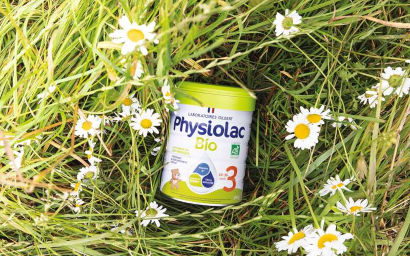 Boîte de lait infantile Physiolac Bio 3 posée dans l'herbe, entourée de fleurs blanches