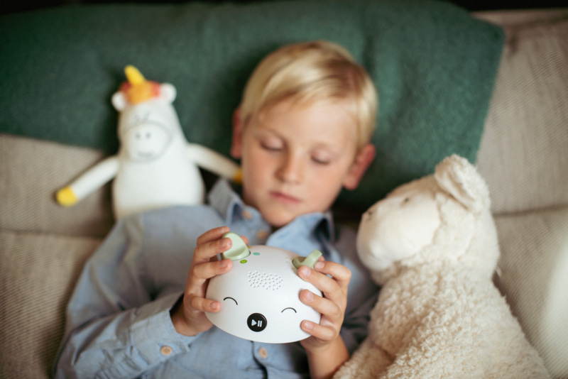 Enfant blond allongé sur un canapé, tenant Elio, une boîte à histoires interactive, entouré de peluches.