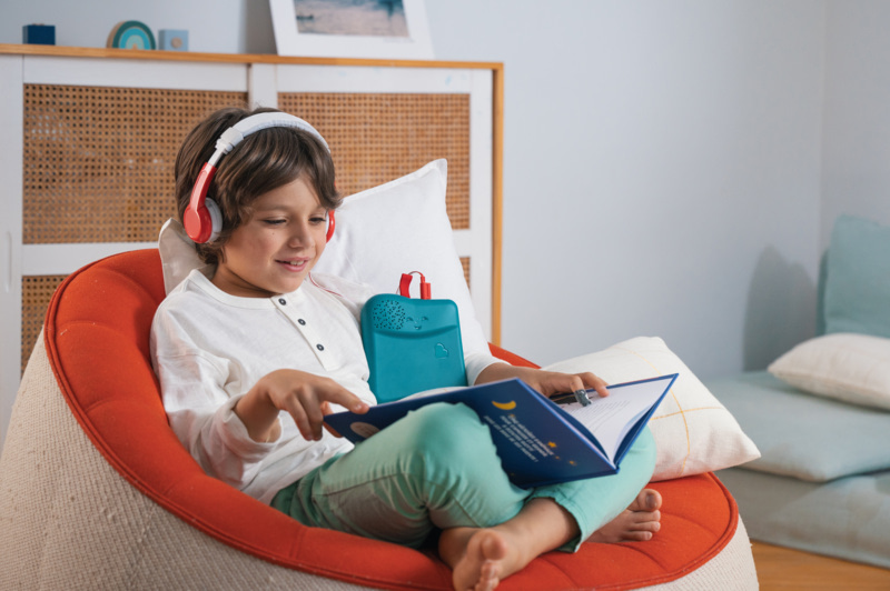 Enfant assis dans un fauteuil orange, portant un casque audio et utilisant Bookinou pour écouter des histoires.